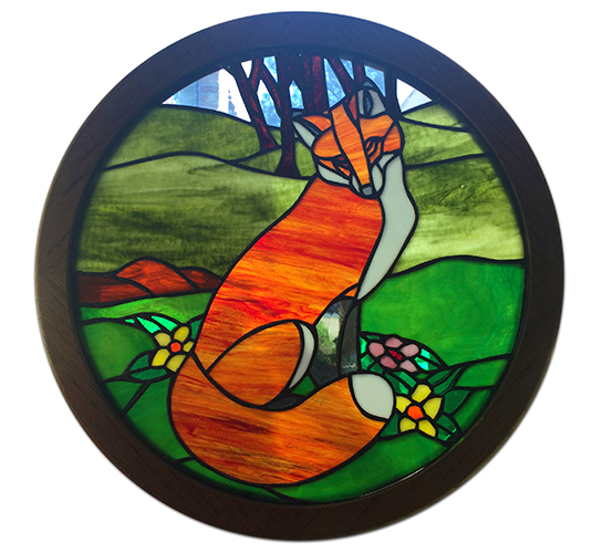 Photograph of the stained glass art depicting Fort Hunt’s fox mascot. An orange, red, and white colored fox is pictured against a green background of rolling hills and trees. The piece is circular in shape and is held in a dark wooden frame.  