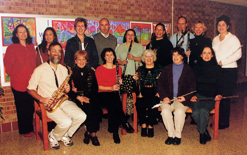 Color photograph from Fort Hunt’s 1999 to 2000 yearbook showing a group of school staff known as the Senior Band. 15 staff members are pictured, most of whom are holding musical instruments. Flutes, clarinets, saxophones, and a trumpet are visible. 